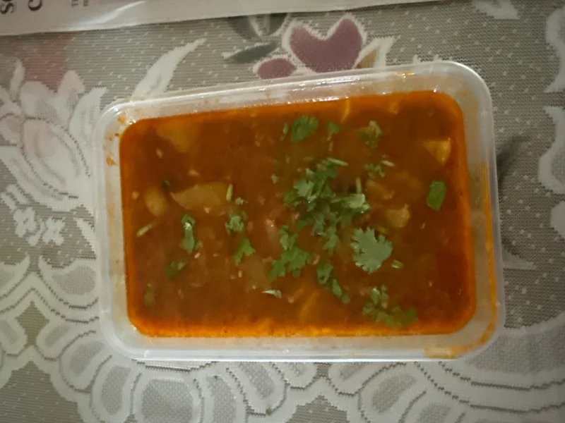 Aloo Rasawala: a thin, red-orange tomato-based curry with potato chunks, garnished with fresh cilantro, served in a rectangular plastic takeaway container.