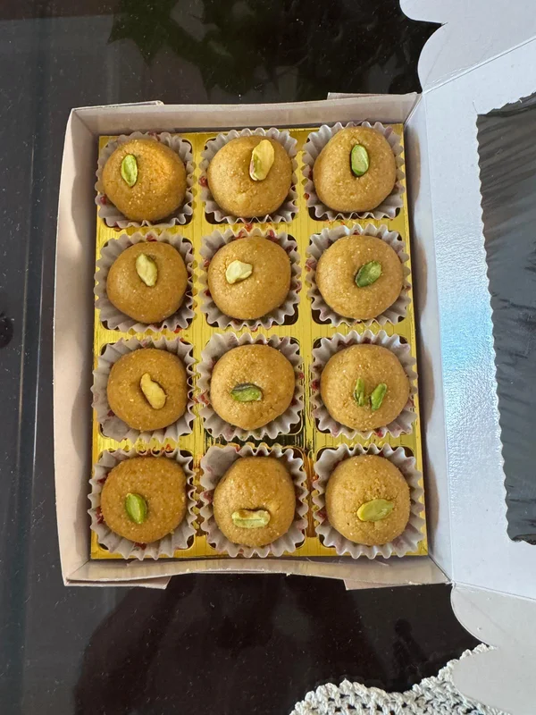 Besan ladoos: twelve round chickpea flour sweets in paper cups, arranged in a box on a gold tray, each topped with a green pistachio.