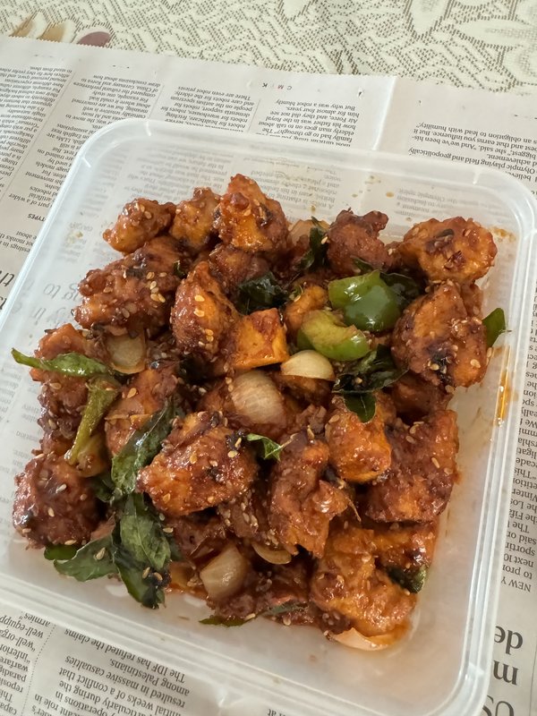Chilli Chicken in a plastic container: fried chicken pieces coated in spicy sauce, tossed with green capsicum, onion, curry leaves, and sesame seeds.