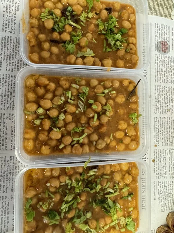 Chole: three plastic containers of chickpea curry in brown spiced gravy, garnished with chopped fresh cilantro, placed on newspaper.