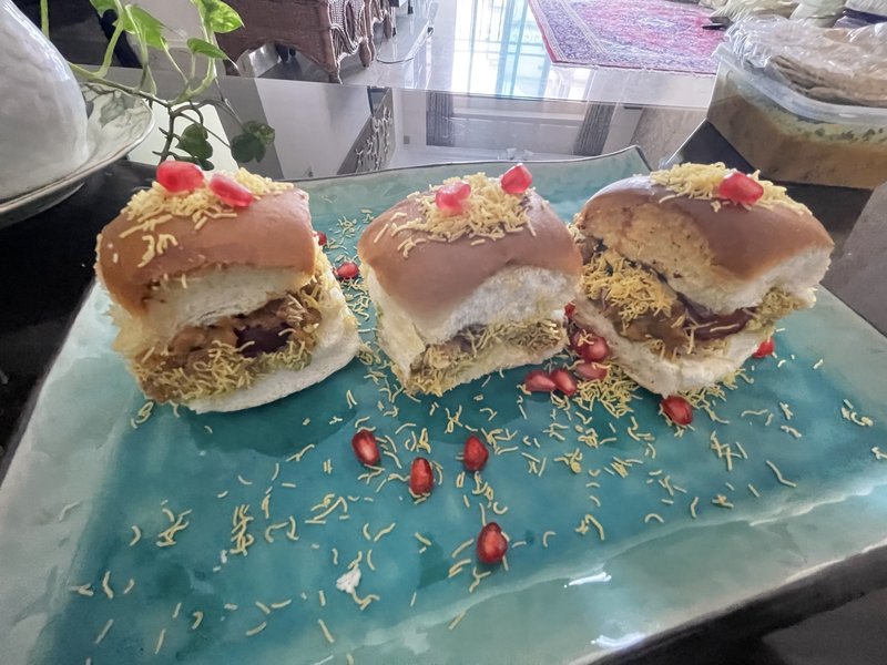 Dabeli: soft buns filled with spiced potato mixture, topped with sev, chopped cilantro, diced onion, pomegranate seeds, and green chutney, served in foil trays.