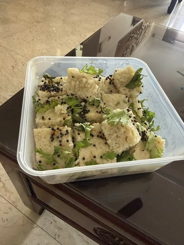 Dhokla Suji (White): steamed semolina cake pieces in a plastic container, topped with black mustard seeds and fresh cilantro leaves.