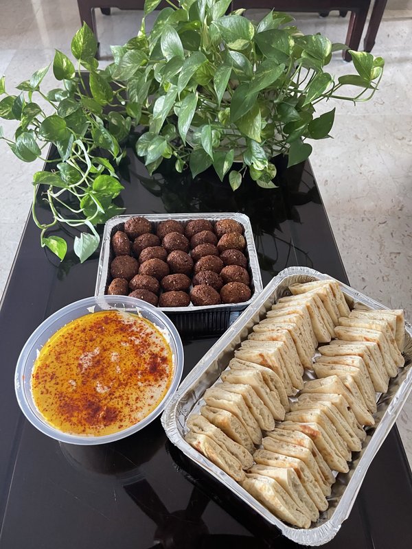 **Pita:** A foil tray filled with approximately 25 round, dark brown fried falafel balls, placed on a black table beside a green pothos plant.