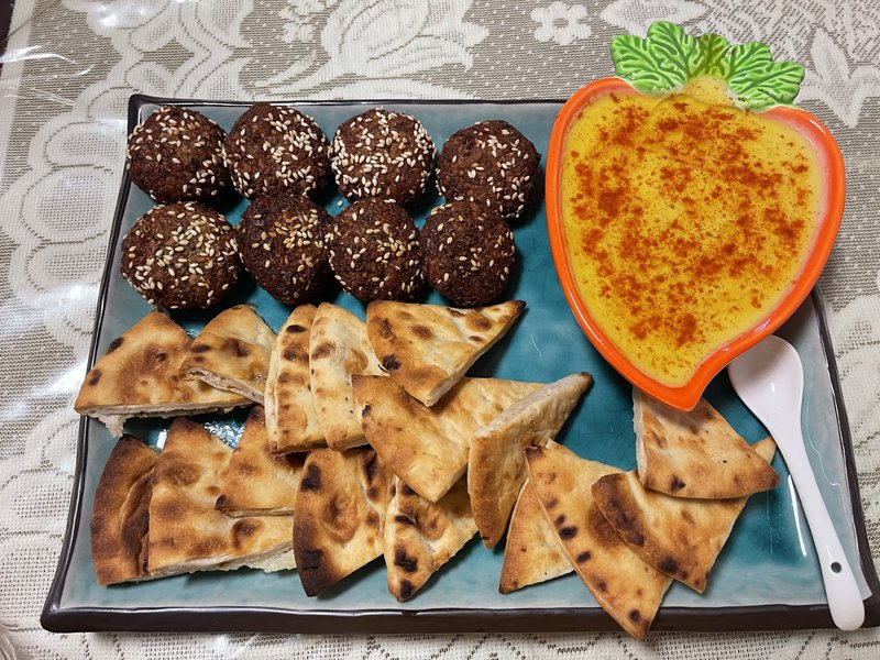 **Hummus (150ml):** A foil tray filled with round, dark brown fried falafel balls, placed on a glossy black table beside a green pothos plant.