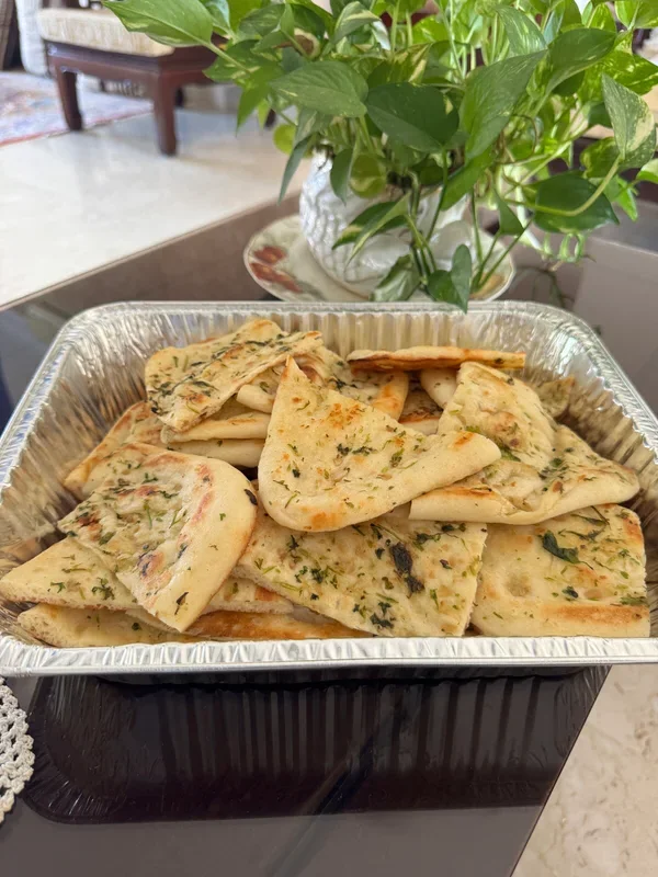 Garlic naan pieces cut into triangles, topped with chopped herbs and garlic, served in a disposable aluminum tray.