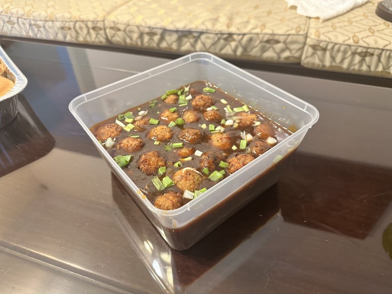 Veg Manchurian: fried vegetable balls in dark brown gravy, topped with chopped green onions, served in a clear plastic takeout container.