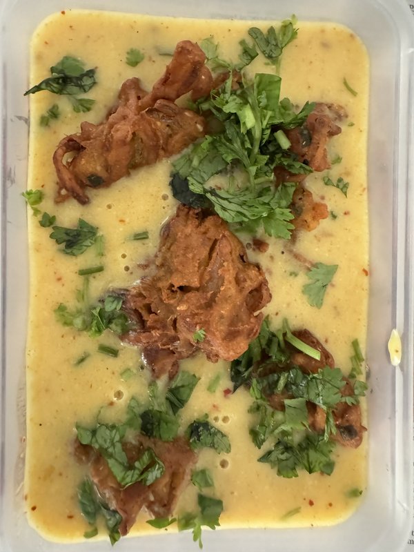 **Kadhi Pakora:** A pot of yellow, turmeric-spiced yogurt-based curry with visible dried red chilies, curry leaves, and a green ladle resting inside.