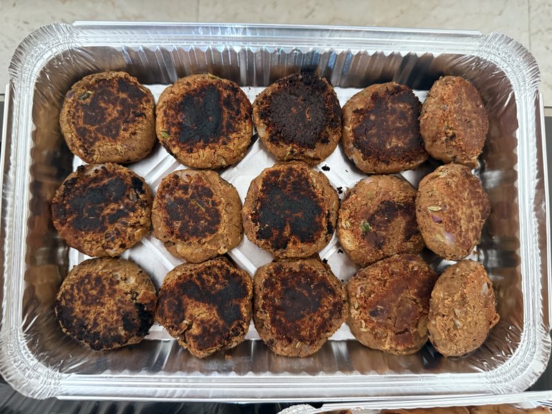 **Veg Galouti Kebab:** Fifteen pan-fried round patties, browned and slightly charred on top, arranged in rows in a rectangular aluminum foil tray, with no garnishes.
