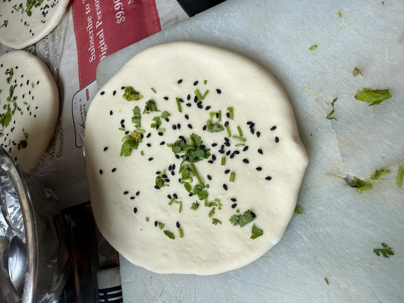 Kulcha: raw flatbread dough topped with chopped cilantro, green chives, and black sesame seeds, resting on a white surface before baking.