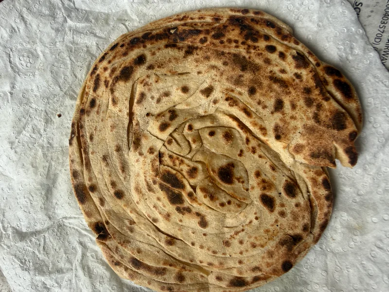 Lachcha Paratha dough rolled flat on a round pan, scored into thin parallel strips dusted with flour, ready to be folded and cooked.