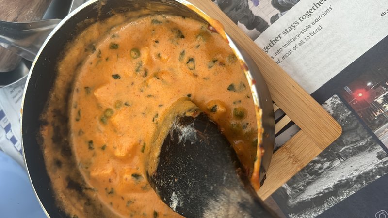 Mattar Paneer: a creamy orange-red curry with cubed paneer and green peas in a pan, stirred with a dark ladle.