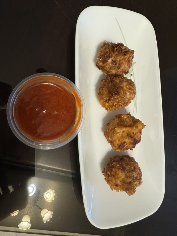 **Veg Momos:** Four round, deep-fried golden-brown dumplings served on a white rectangular plate with a small cup of red chili dipping sauce.