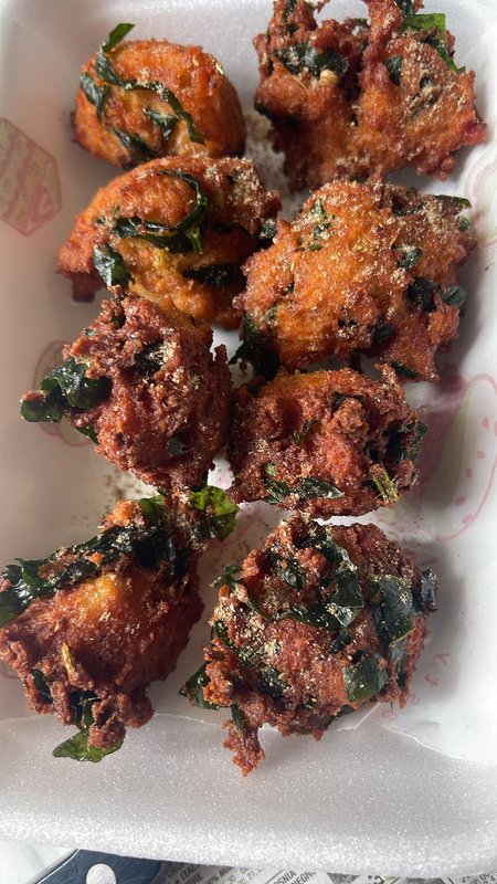 Onion Pakora: deep-fried battered fritters with visible onion and chopped curry leaves, coated in reddish spiced batter, served in a styrofoam container.