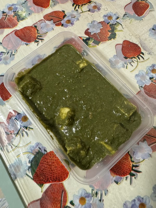 Palak paneer in a plastic takeaway container: a thick dark-green spinach sauce with visible cubes of white paneer cheese.
