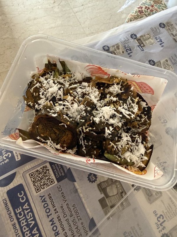 Patra served in a takeout container, consisting of coiled taro leaf rolls topped with shredded coconut, sesame seeds, and curry leaves.
