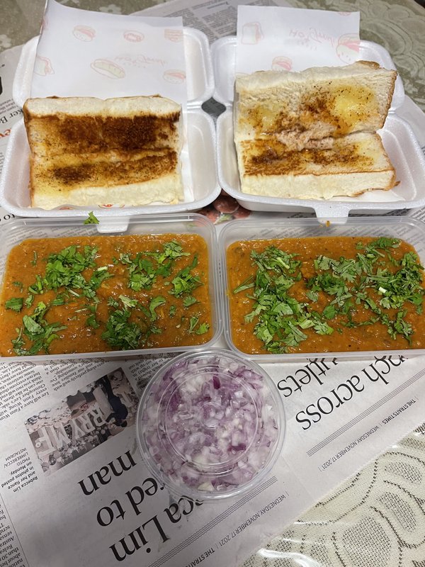 **Pav Bhaji:** Two takeout containers of spiced mashed vegetable curry garnished with chopped cilantro, served with toasted bread rolls and a side of diced raw onions.