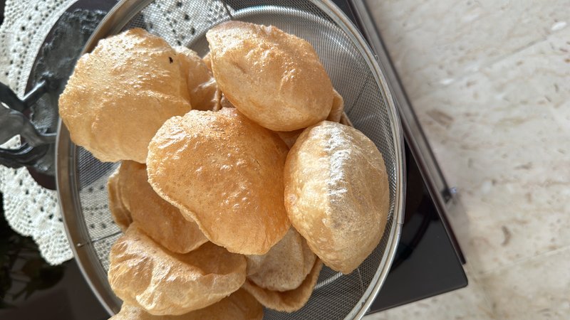Puri (Plain): deep-fried, puffed whole-wheat bread rounds, golden-brown in colour, stacked in a steel mesh strainer/colander.
