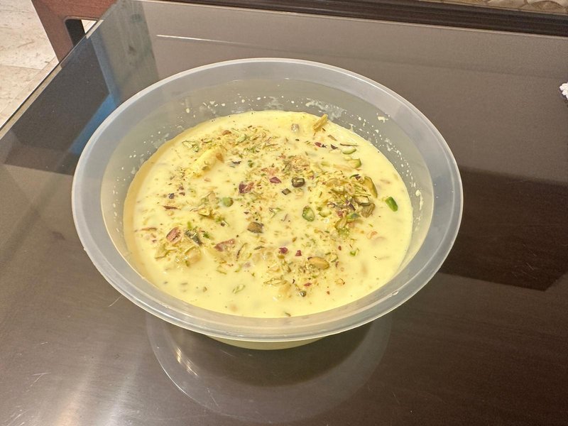 Rasmalai served in a clear plastic bowl, consisting of a creamy yellow milk sauce topped with chopped pistachios and mixed nuts.