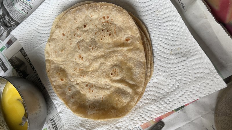 Wheat Roti: a stack of cooked flatbreads on paper towels beside a single roti cooking on a flat black tawa griddle on a stovetop.