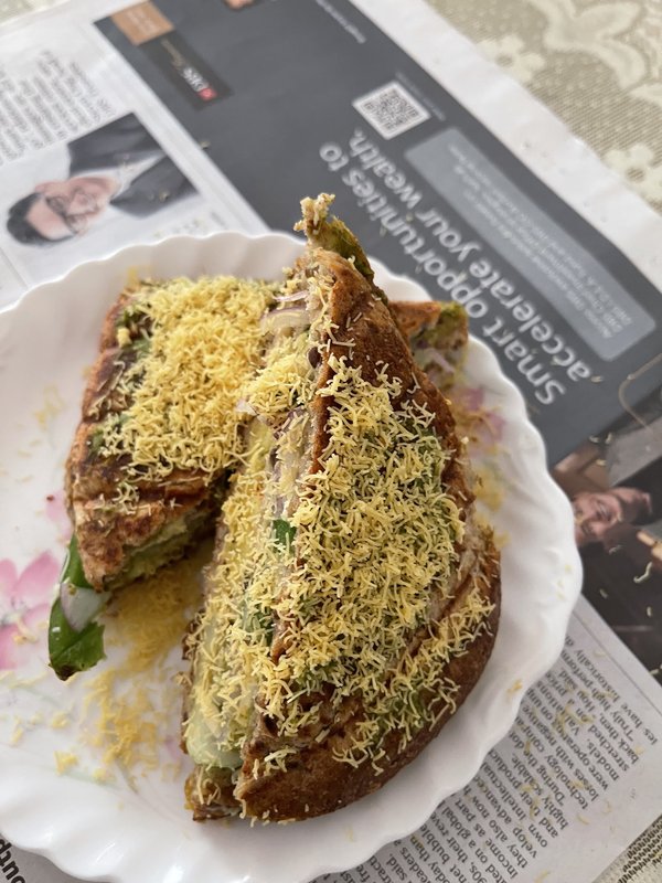 Mumbai Cheese Toasted Sandwich: two toasted brown bread triangles topped with grated yellow sev, filled with green chutney and vegetables, served on a white scalloped plate.