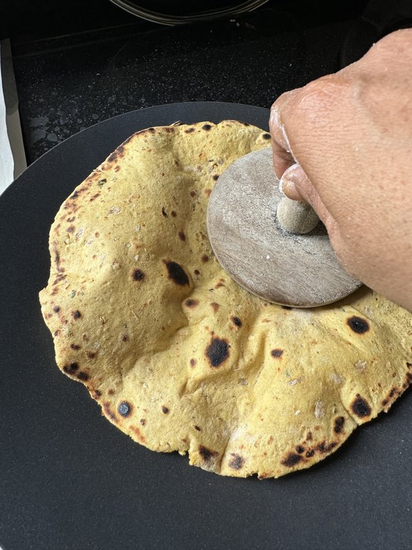 **Thepla (Dal):** A thin, round, rolled-out flatbread dough made with lentils, spiced with visible red chili flakes and green herbs, resting on a metal rolling surface dusted with flour.