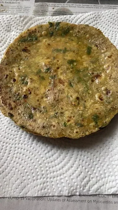 Thepla (Methi): a large, round, uncooked flatbread on a pan, flecked with green fenugreek leaves, with a cooked thepla visible underneath.