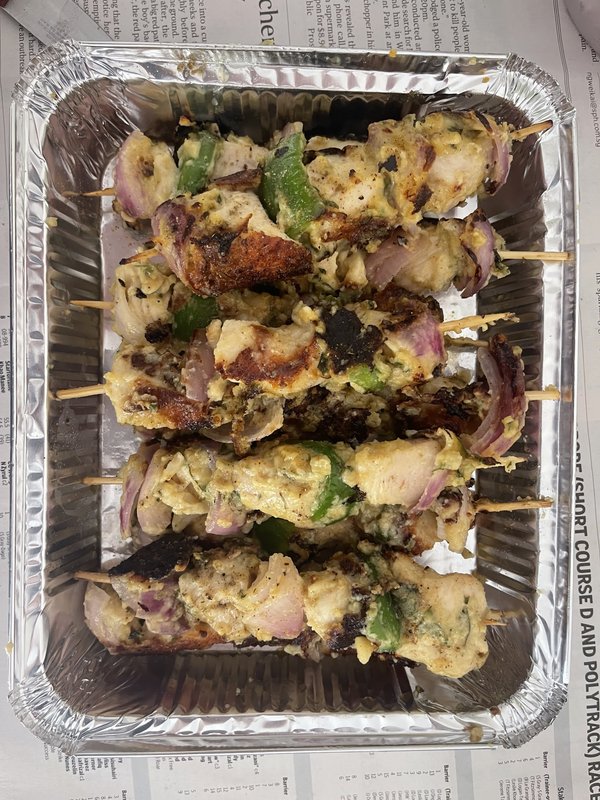 Malai Chicken Tikka: grilled chicken chunks on wooden skewers with red onion and green bell pepper pieces, served in a foil tray lined with newspaper.