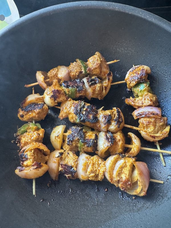 Chicken Tikka skewers coated in red-orange spiced marinade, with visible onion and green pepper pieces, served on a glass plate atop newspaper.