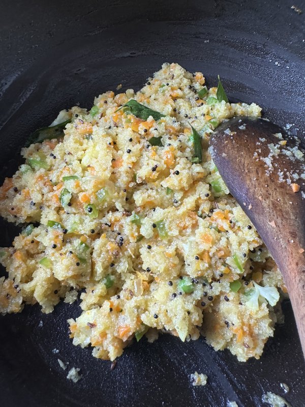 Upma cooked in a black wok with semolina, diced carrots, green vegetables, mustard seeds, and curry leaves, being stirred with a wooden spatula.