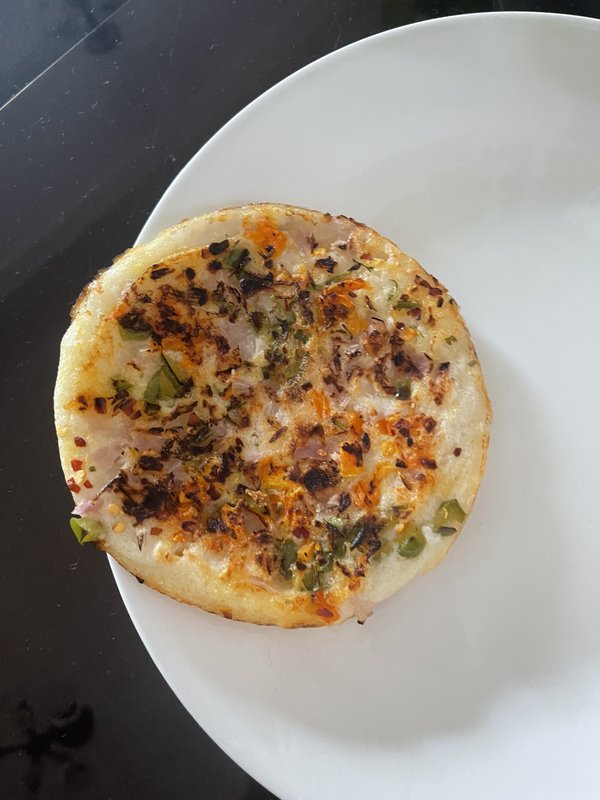 Mini Uttapam on a white plate, topped with chopped green peppers, red onion, melted cheese, red chili flakes, and herbs, with browned, slightly charred edges.