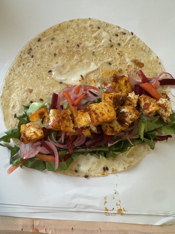 **Tandoori Paneer Wrap** — a round flatbread with seeds, spread with a layer of white cream cheese or yogurt-based sauce, laid flat on white paper.